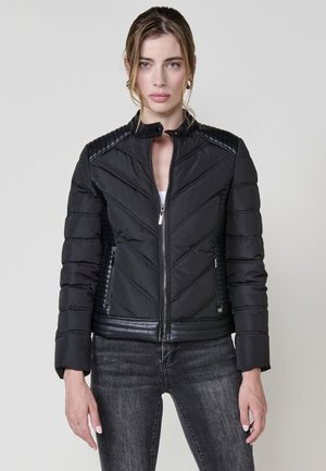 Chaqueta acolchada negra con acolchado en forma de chevron, cuello elevado, cierre de cremallera y detalles en cuero sintético negro en los hombros y el dobladillo.