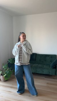 Mladá žena s copom má na sebe sivú mikinu s kapucňou, pruhované tričko a široké džínsy, stojí v jasnej miestnosti so zelenou sedačkou a izbovou rastlinou v kvetináči.