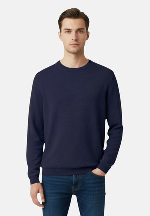 Uomo in piedi, di fronte, che indossa un maglione girocollo blu scuro e jeans blu, con una mano in tasca, su uno sfondo bianco neutro.