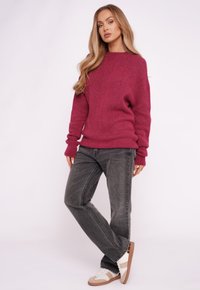 Bordowy sweter z wełny o luźnym kroju, z długimi rękawami i ściągaczami, zestawiony z szarymi, wysokim stanem dżinsami oraz białymi tenisówkami z brązowymi akcentami.