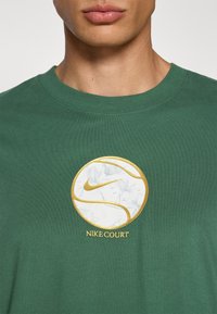 Grøn bomulds-T-shirt med et guld- og hvidt rundt logo med blomstermønster og teksten "NIKE COURT" nederst. Klassisk pasform.