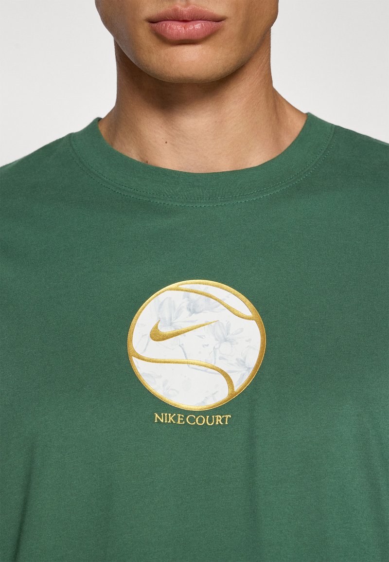 T-shirt verde in cotone con un logo circolare oro e bianco dotato di un motivo floreale e la scritta "NIKE COURT" in basso. Vestibilità classica.
