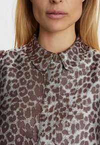 Blusa in stampa animale marrone con una leggera lucentezza, collo a camicia e un fermaglio a forma di cuore in metallo sul davanti.