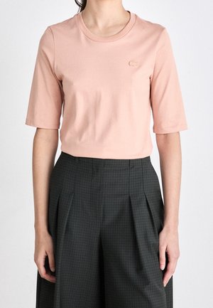 Vrouw in een perzikkleurig shirt met korte mouwen, gestopt in een donkergrijze hoge taille plooibroek met een subtiel ruitpatroon.