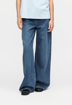 Personne portant une chemise bleu clair et un jean large bleu ample couvrant des chaussures noires brillantes, debout devant un fond blanc.