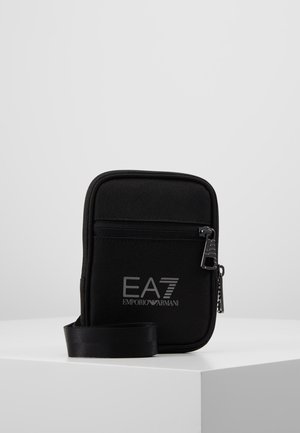 Czarna kompaktowa torba typu crossbody z przednią kieszenią na zamek błyskawiczny i logo EA7 Emporio Armani, prezentowana na białym pedestale.