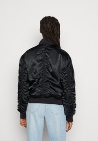 Giacca bomber in satin nera con dettagli arricciati lungo le maniche e il retro. Presenta un orlo e polsini a costine, abbinata a jeans azzurri chiari.