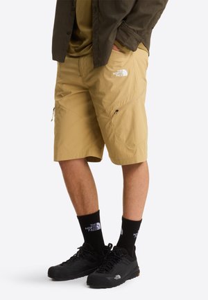 Kaki korte broek van lichtgewicht stof, voorzien van twee ritssluitingen en een logopatch. Gecombineerd met zwarte schoenen en sokken.