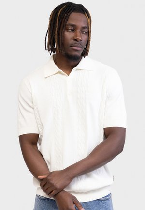 Νέος άνδρας με κοντά ντρέντλοκς που φοράει λευκό πικέ πόλο με κοντά μανίκια και ανοιχτόχρωμο τζιν, στέκεται μπροστά σε έναν απλό φόντο.