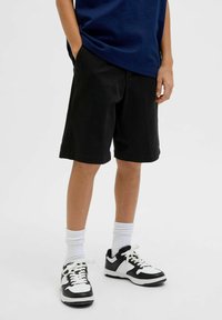 Shorts en coton noir avec une coupe droite, des poches latérales et une coupe décontractée. Associés à des chaussettes de sport blanches et des baskets noires et blanches.