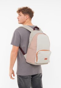 Batoh v béžovo-ružovej farebnej kombinácii, s textúrovaným povrchom, predným zipsovým vreckom a burgundskými popruhmi.