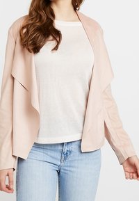 Saskia rosa öppen jacka i mjukt material, med draperade flikar och långa ärmar, lagd över en ljus beige topp.