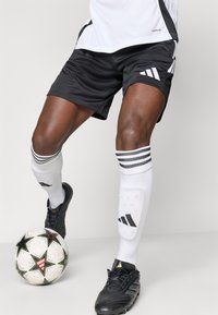 Μαύρα αθλητικά σορτσάκια με λευκές λεπτομέρειες, συνδυασμένα με μαύρες ποδοσφαιρικές μπότες και λευκές κάλτσες με μαύρες ρίγες. Ένας κανονισμός ποδοσφαίρου είναι ορατός.