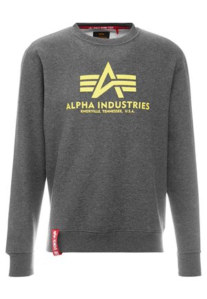 Grauer Pullover aus weichem Material mit einem großen gelben "Alpha Industries" Logo mit Streifen und einem roten Tab am Saum.