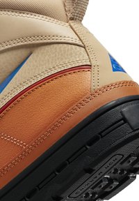 Tan leather sneaker with blue and red accents, textured side panels, and a black rubber sole featuring tread for grip.