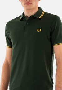 Groene polo met een gestructureerde stof, voorzien van een drieknoopssluiting, een kraag en manchetten met gele strepen, en een klein geel logo op de borst.