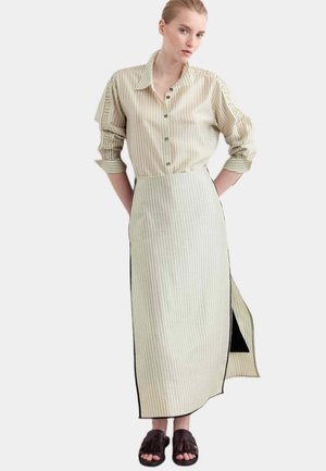 Donna che indossa una camicia beige a righe con bottoni e maniche arrotolate e una gonna lunga abbinata con spacco laterale, abbinata a sandali con nappine marrone scuro.