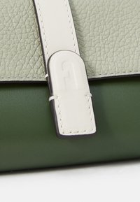 Sac à main en cuir vert avec une bandoulière texturée vert clair, un accent blanc et un logo en relief. Coutures apparentes.