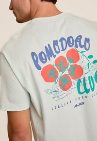 Lichtgroen katoenen t-shirt met een grote graphic van rode tomaten, groene bladeren en blauwe tekst met "POMODORO" en Italian Food Club.