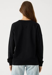 Sudadera negra con cuello redondo y mangas largas. Presenta un corte relajado y una textura suave. Combinada con vaqueros de mezclilla de color azul claro.