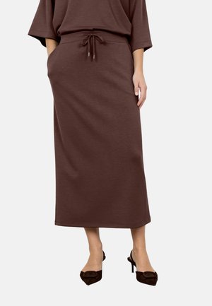 Woman wearing a brown midi skirt with a drawstring waist and matching brown heeled shoes, hands resting in pockets.