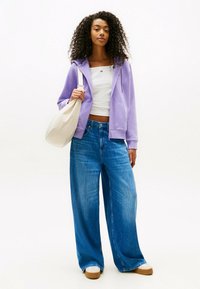 Hellblaues ZIP-Hoodie über einem weißen Croptop, kombiniert mit weit geschnittenen blauen Jeans. Hält eine cremefarbene Tragetasche. Holzboden-Schuhe.