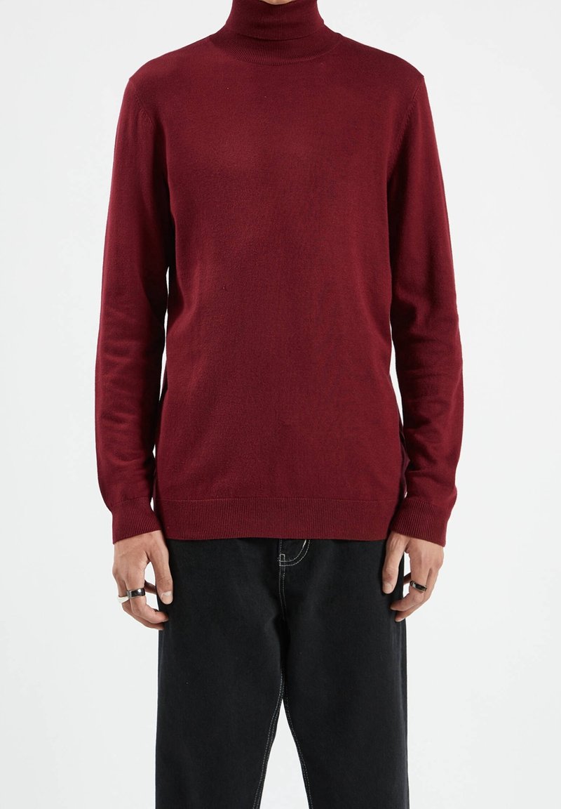 Personne portant un pull à col roulé bordeaux à manches longues et un pantalon noir, avec des accessoires en forme d'anneaux aux deux mains.