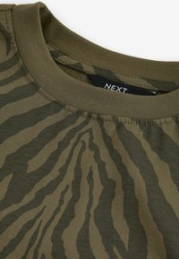 Koszulka w oliwkowym kolorze z okrągłym dekoltem, z ciemnozielonym wzorem w paski zebry i czarną metką z białym napisem "NEXT" wewnątrz kołnierza.