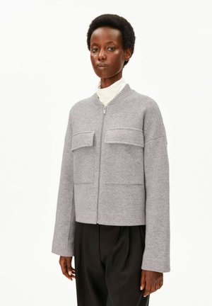 Frau, die eine graue Jacke mit Reißverschluss und Klappentaschen über einem weißen Rollkragenpullover und schwarzen Hosen trägt, steht vor einem einfarbigen weißen Hintergrund.