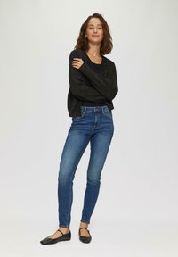 Schwarze gemusterte Strickjacke über einem schwarzen eng anliegenden Oberteil, kombiniert mit mittelblauen Skinny-Jeans und schwarzen Ballerinas mit Knöchelriemen. Schlichter Hintergrund.