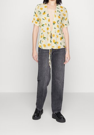 Gul skjorte med knapper og print af citroner og blade, løs pasform, og korte ærmer, parret med brede mørke jeans og sorte platform-sko.