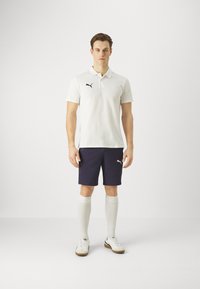 Bílý polo triko s černým logem, námořnické šortky, bílé ponožky po kolena a bílé sportovní boty s béžovými detaily. Jednoduchý a sportovní outfit.