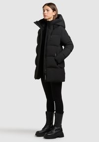 Parka nera imbottita con cappuccio, dotata di zip frontale, tasche laterali e sezioni trapuntate. Abbinata a stivali cargo neri con stringhe.