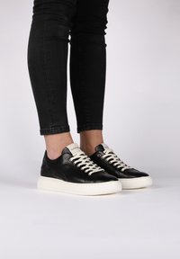 Černé kožené tenisky s bílou gumovou podrážkou, se smetanovými tkaničkami a béžovou štítků značky na jazyku, zobrazené na nohou s černými džíny.