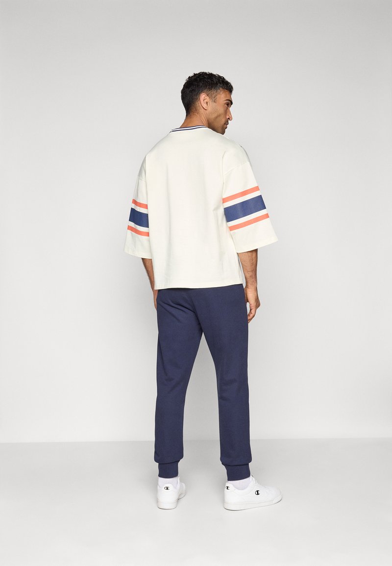 Lichtcrème oversized shirt met gestreepte mouwen in blauw, rood en wit, gecombineerd met marineblauwe joggingbroek en witte sneakers. Huidige stoftextuur.