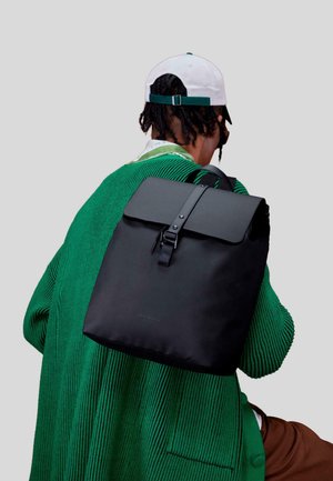 Zaino nero con chiusura a patta, tracolla regolabile e dettaglio in fibbia. Indossato sopra un maglione verde a coste, con tessuto testurizzato.