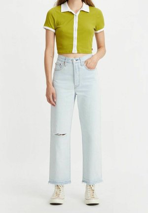 Woman wearing light blue ripped jeans, white sneakers, and a cropped green polo shirt with white collar and trim.