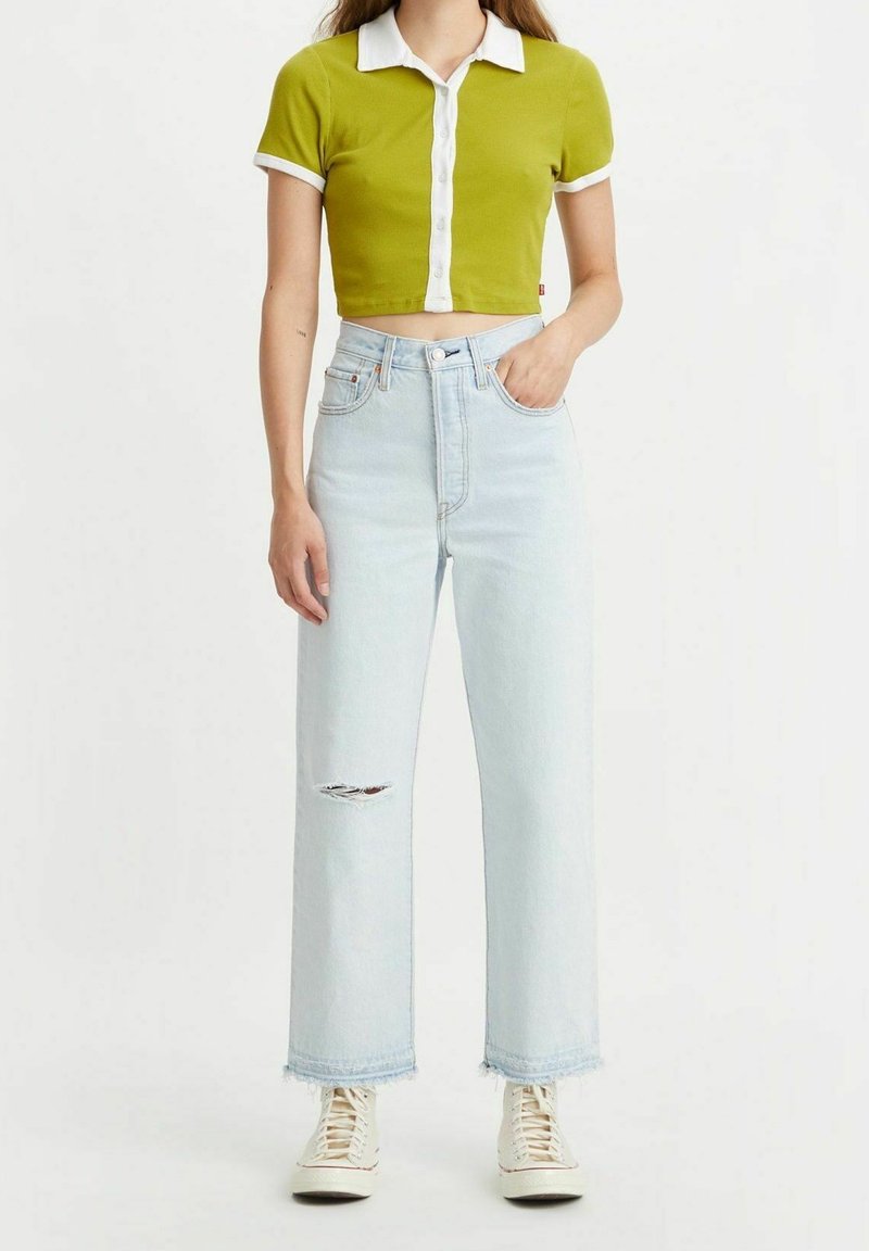 Femme portant un jean bleu clair déchiré, des baskets blanches et un polo vert court avec un col et des bordures blanches.
