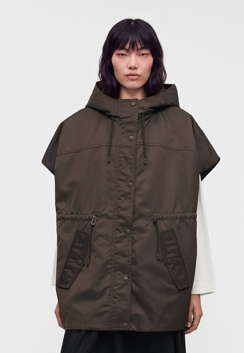 Personne portant une veste sans manches oversize à capuche vert foncé avec des boutons devant et de grandes poches à rabat sur un haut blanc à manches longues.