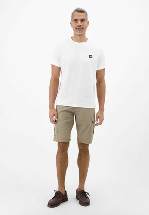 Mand stående iført hvid kortærmet t-shirt med logo, beige cargoshorts og brune snørebåndssko på hvid baggrund.