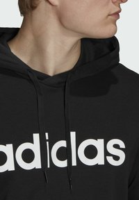 Felpa con cappuccio nera caratterizzata da un vistoso logo "adidas" bianco. Il tessuto è liscio con una vestibilità standard e presenta lacci regolabili.
