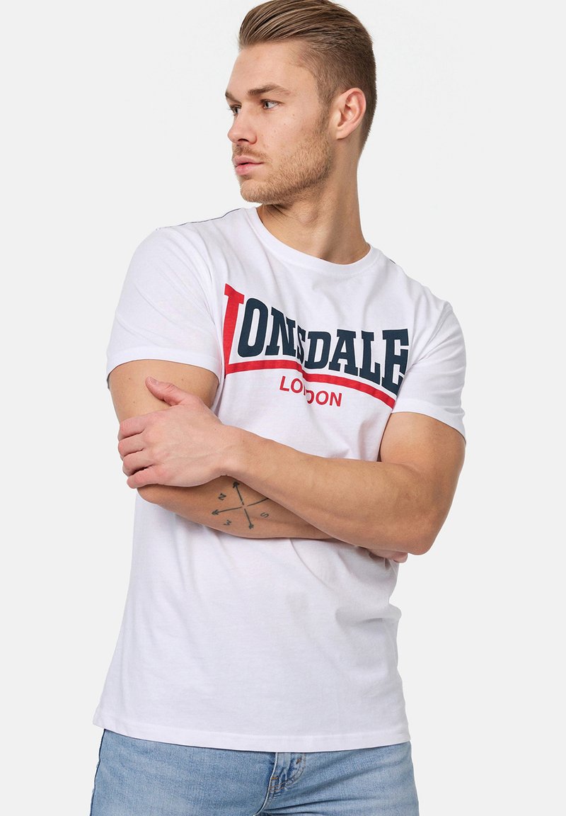 Bjela pamučna majica kratkih rukava s odvažnim crno-crvenim logom "LONSDALE". Klasičan okrugli izrez i kratki rukavi. Nosena s svijetloplavim trapericama.
