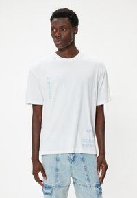 Vit bomull t-shirt med vertikala blå textgrafik. Avslappnad passform med korta ärmar och rundad krage. Slät textur.