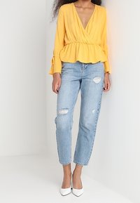 Blusa gialla a peplo con scollo a V profondo e maniche lunghe, abbinata a jeans azzurri scoloriti e tacchi bianchi a punta.