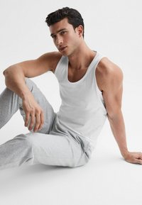 Vit ärmlös tanktop tillsammans med ljusgråa sweatpants, med en avslappnad passform och mjuk bomullstextur. Modellen sitter mot en vit bakgrund.