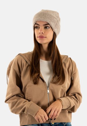 Jeune femme portant un bonnet en tricot beige et une veste zippée marron, regardant légèrement sur le côté, les mains jointes devant elle.