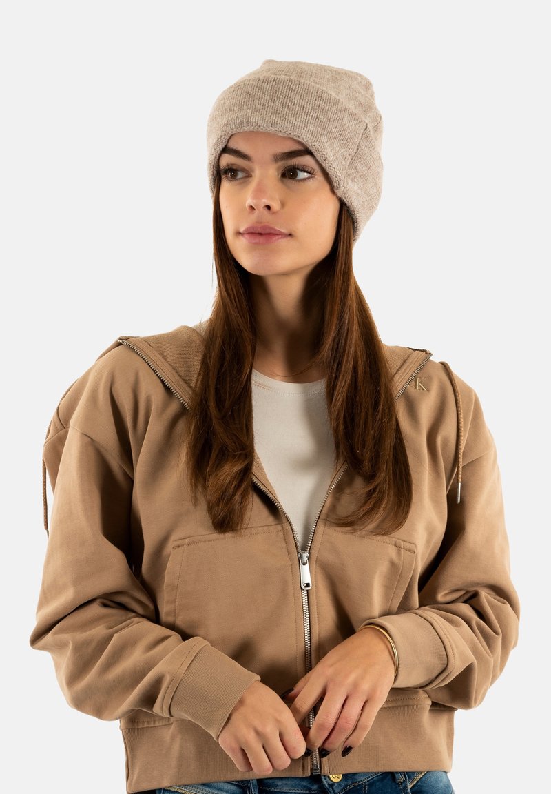 Jeune femme portant un bonnet en tricot beige et une veste zippée marron, regardant légèrement sur le côté, les mains jointes devant elle.