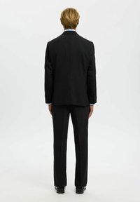 Zwarte op maat gemaakte kostuum met een jasje met een enkele split, vlakke broek en een lichtblauwe shirt. Gladde stof en gestructureerd ontwerp.