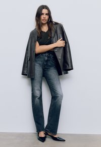 Črna usnjena oversize jakna, temno zelena majica, zbledeli ravni kavbojke in črne konične pete. Gladke teksture in slojevita zasnova.