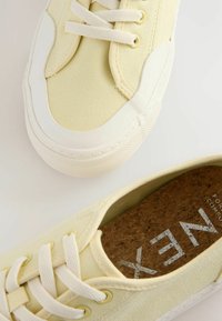 Baskets en toile jaune clair avec semelles en caoutchouc blanc, présentant des lacets ronds et une semelle intérieure en liège texturé avec le nom de la marque imprimé.